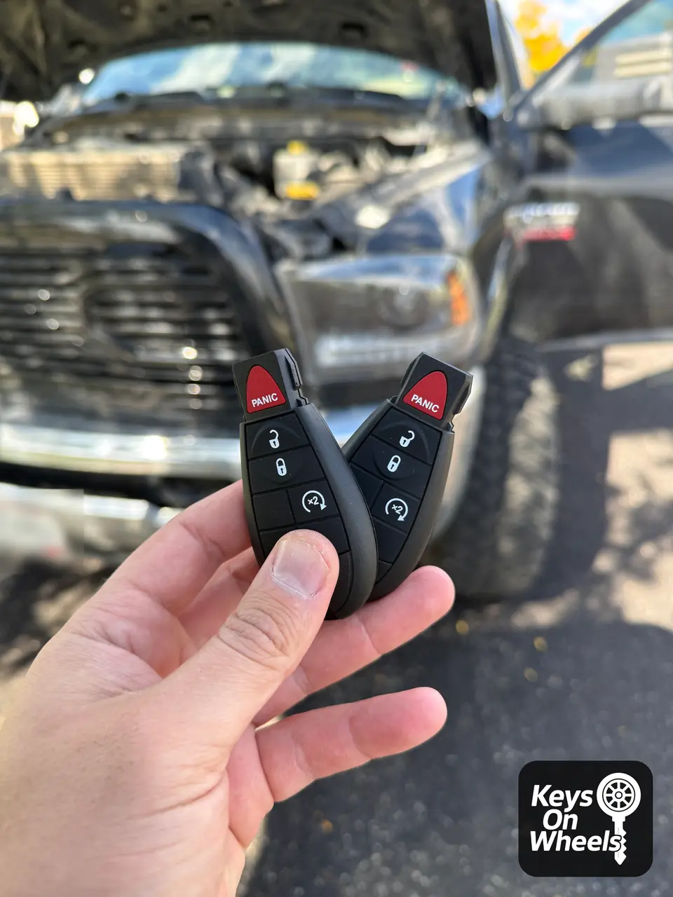 Two fobik keys with remote start and panic buttons, held in front of a pickup with its hood open in Meridian